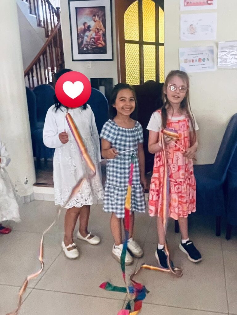 Three girls in dresses holding rainbow ribbons by the front door of the home
