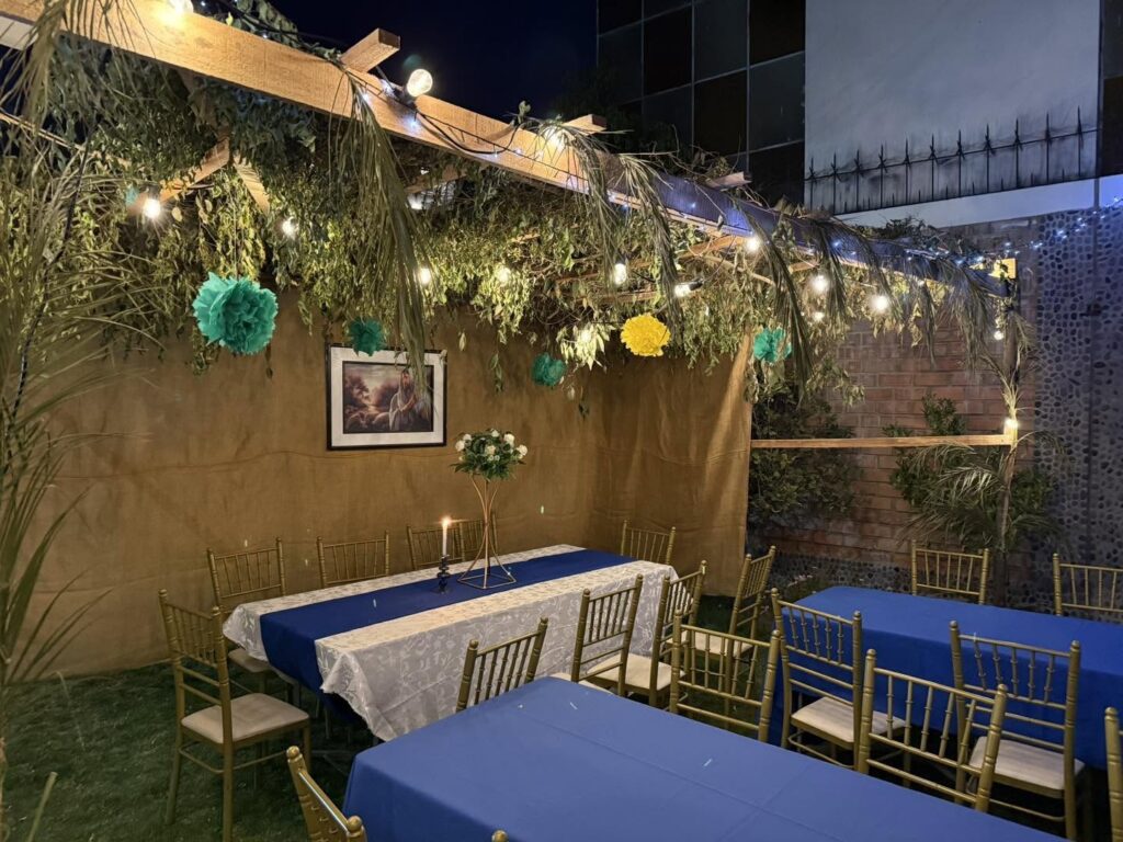 The sukkah lit with string lights at night, tables set in blue and white with a single candle